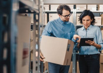 Female Inventory Manager Shows Digital Tablet Information to a Worker Holding Cardboard Box, They Talk and Do Work. In the Background Stock of Parcels with Products Ready for Shipment.