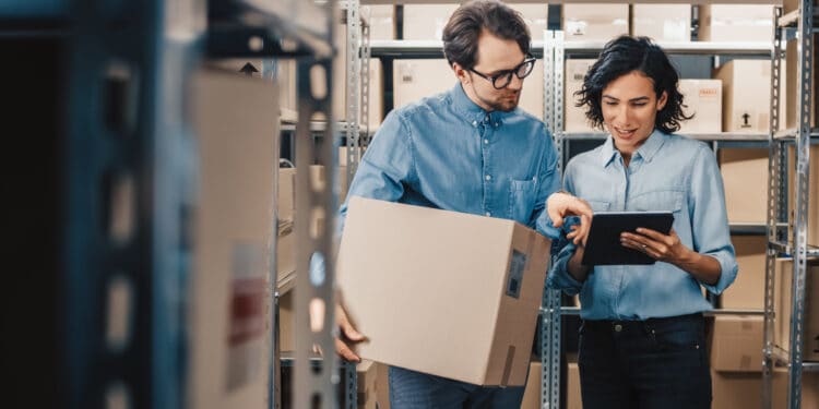 Female Inventory Manager Shows Digital Tablet Information to a Worker Holding Cardboard Box, They Talk and Do Work. In the Background Stock of Parcels with Products Ready for Shipment.